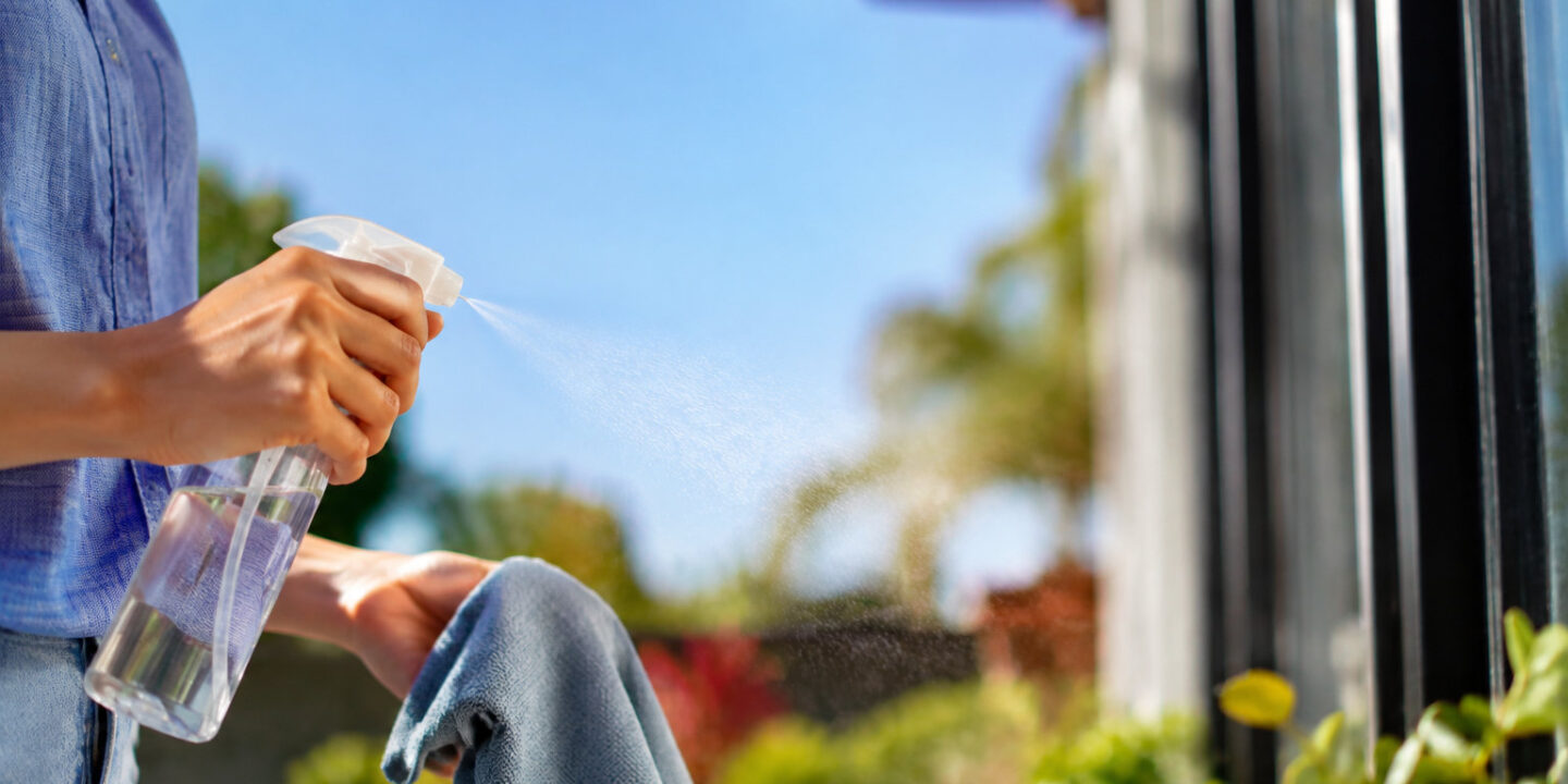 Une personne vaporise un produit nettoyant sur une vitre avec un chiffon en main, avec un jardin en arrière-plan.